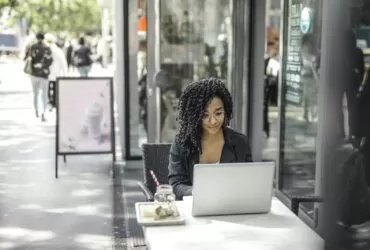 High angle of pensive African American female freelancer in glasses and casual clothes focusing on screen and interacting with netbook while sitting at table with glass of yummy drink on cafe terrace in sunny day
